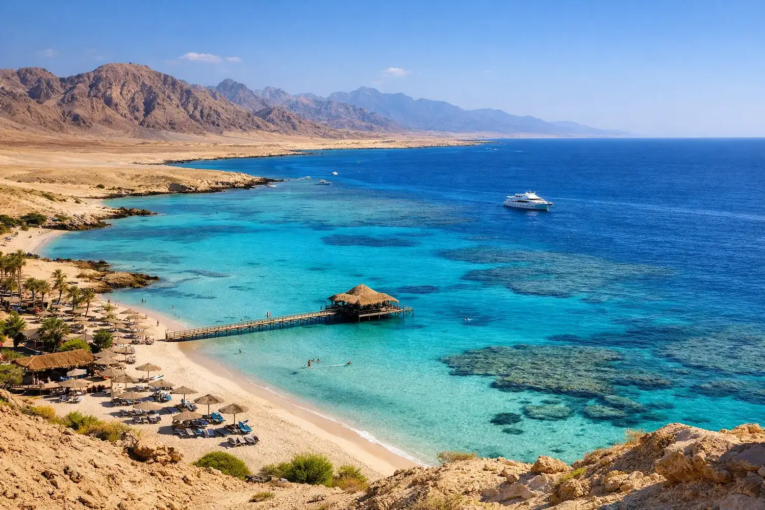 Discover the Best Things to do in Marsa Alam. Aerial view of Marsa Alam coastline with turquoise Red Sea water, sandy beach, wooden pier, coral reefs, and desert mountains